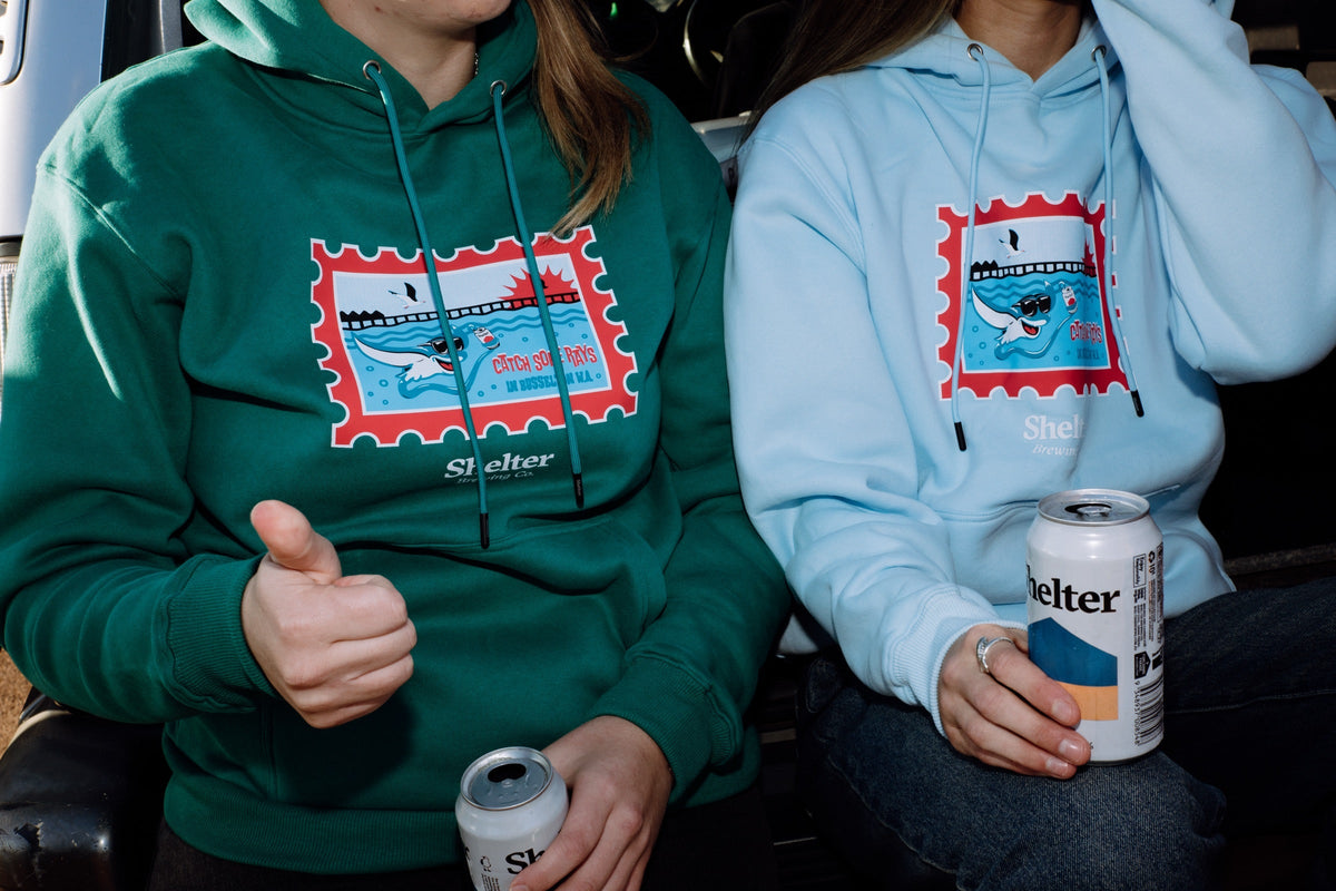 Two Shelter hoodies in green and pale blue, each with a red screen printed stamp graphic on the chest