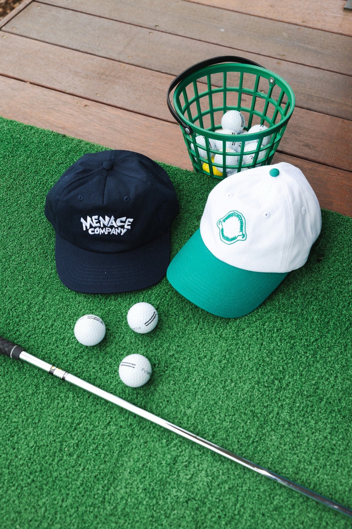 Two Menace Company embroidered caps in navy and white with a green brim, laid on a putting green with golf balls