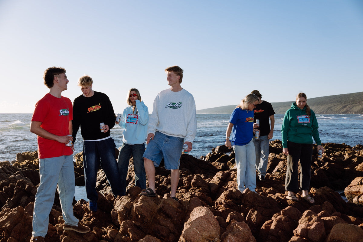 Seven people in the Shelter printed tee, crew and hoodie range on the rocks above a beach at sunset